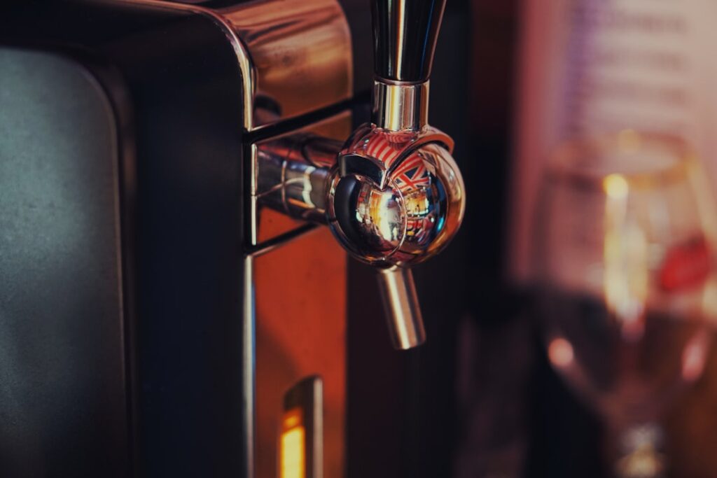 Person stirring malt extract into a large stainless steel pot on a kitchen stove with brewing supplies nearby.