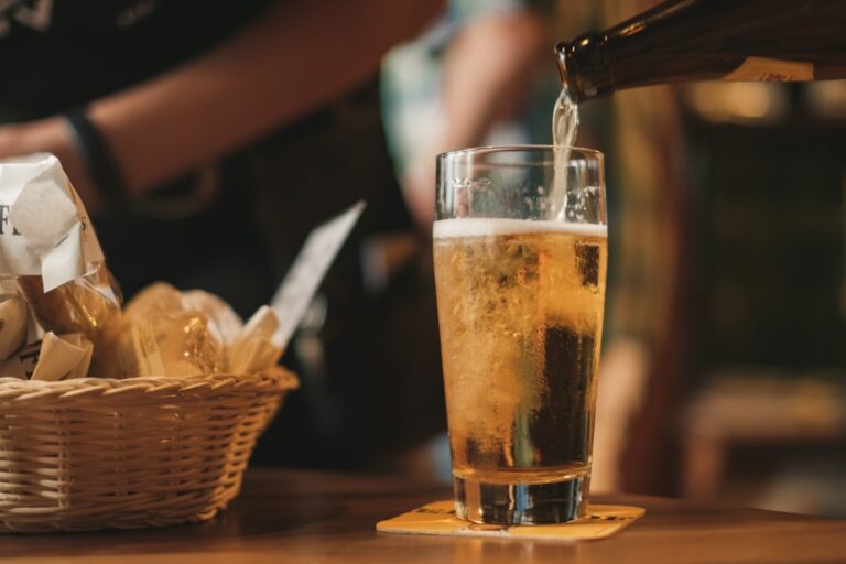 A person pouring beer from a bottle into a tilted pint glass on a wooden table indoors.