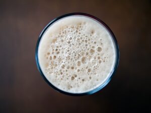 A close-up of a hand holding a pint glass with golden beer, foam head, and visible hop cones in the background.