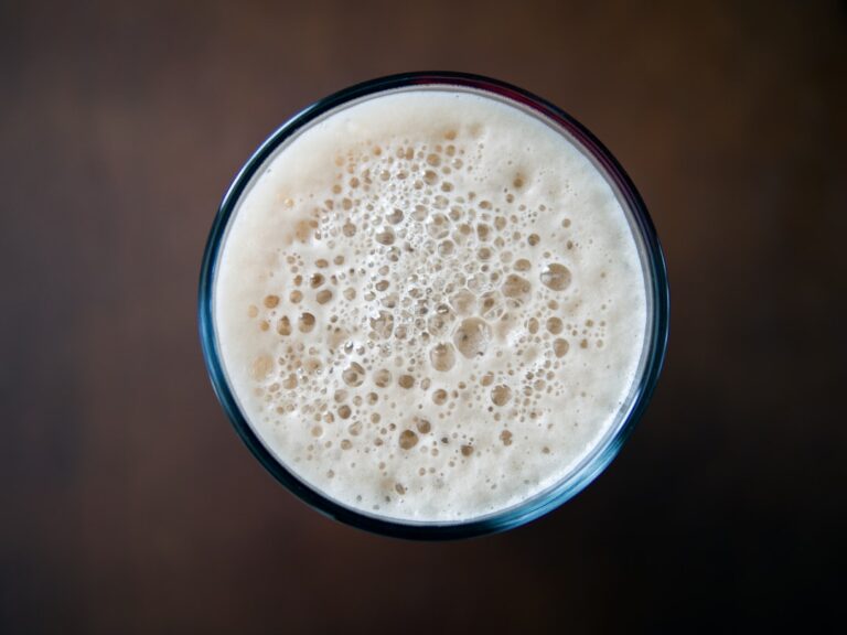 A close-up of a hand holding a pint glass with golden beer, foam head, and visible hop cones in the background.