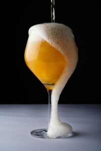 A person pours a hazy, golden sour beer into a tulip glass on a wooden table.