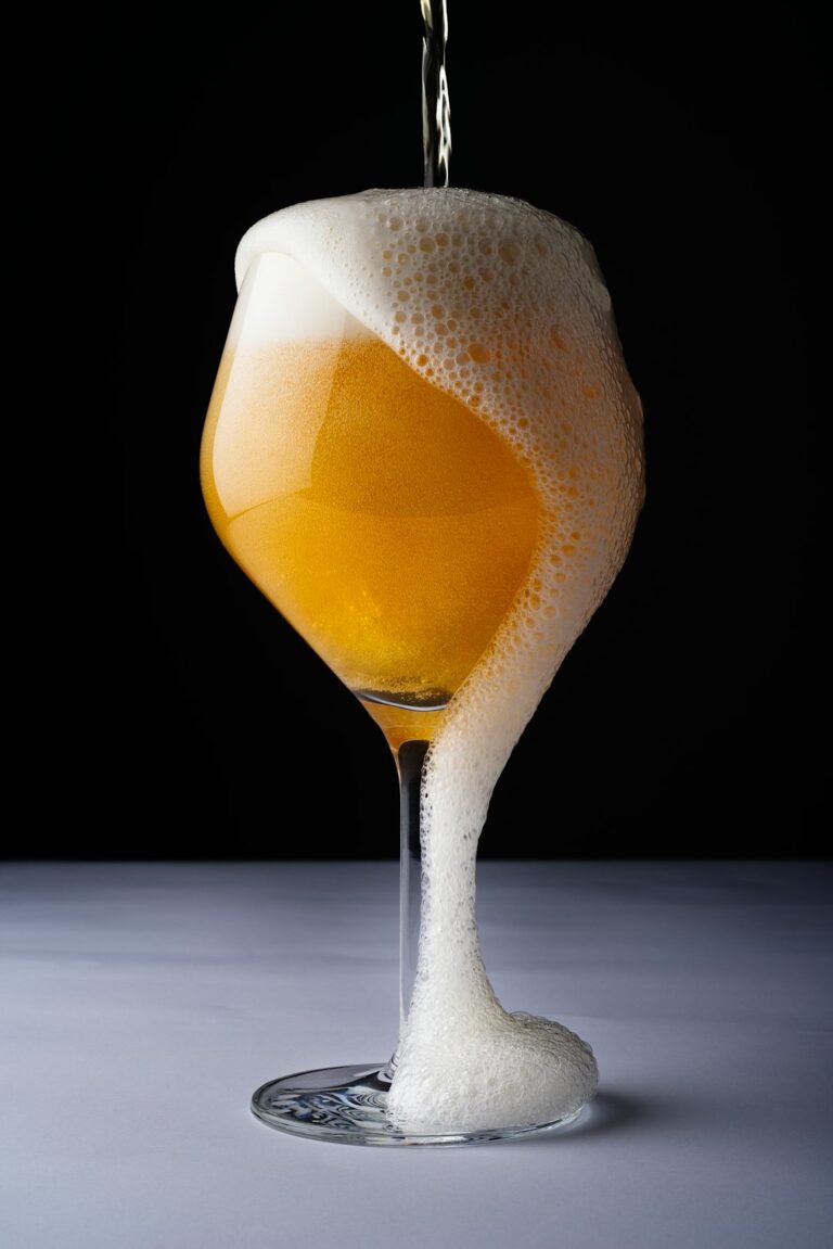 A person pours a hazy, golden sour beer into a tulip glass on a wooden table.