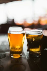 Two glasses of pale beer sit beside hops and malt on a wooden tasting table.